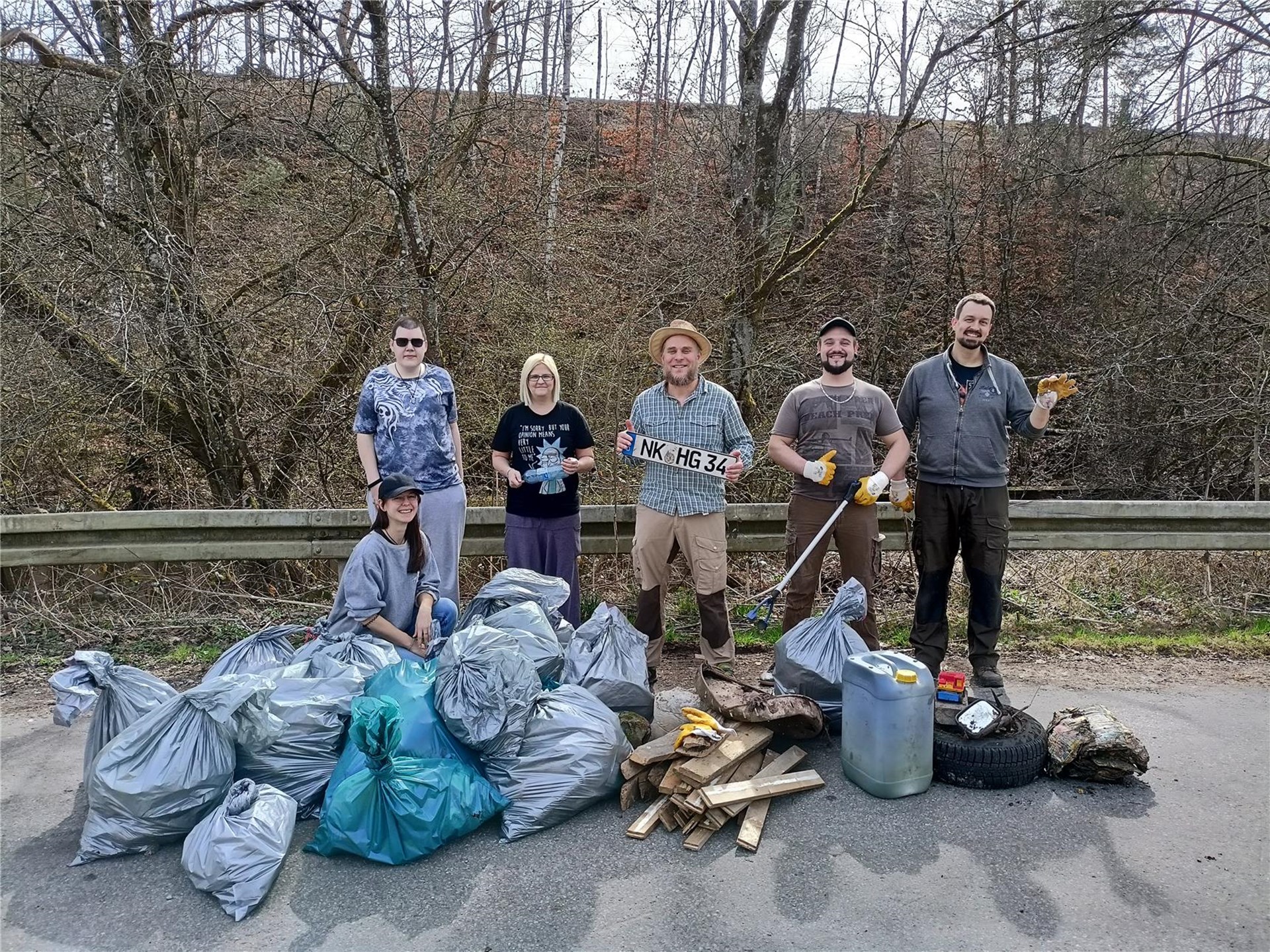 Mitarbeitende des Teams stehen zusammen mit Klientinnen und Klienten hinter mehreren Mülltüten. Sie halten dicke Handschuhe und Müllsammler in den Händen, außerdem ein gefundenes Nummernschild  