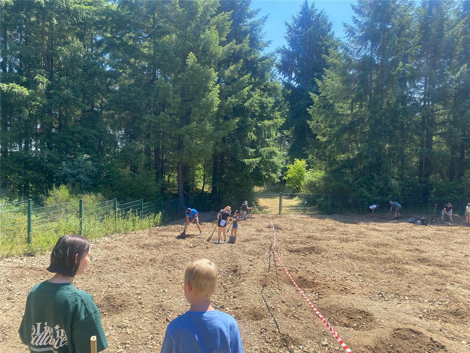 2 Kinder sind von hinten auf einer Sandfläche zu sehen. Im Hintergrund sind Bäume.