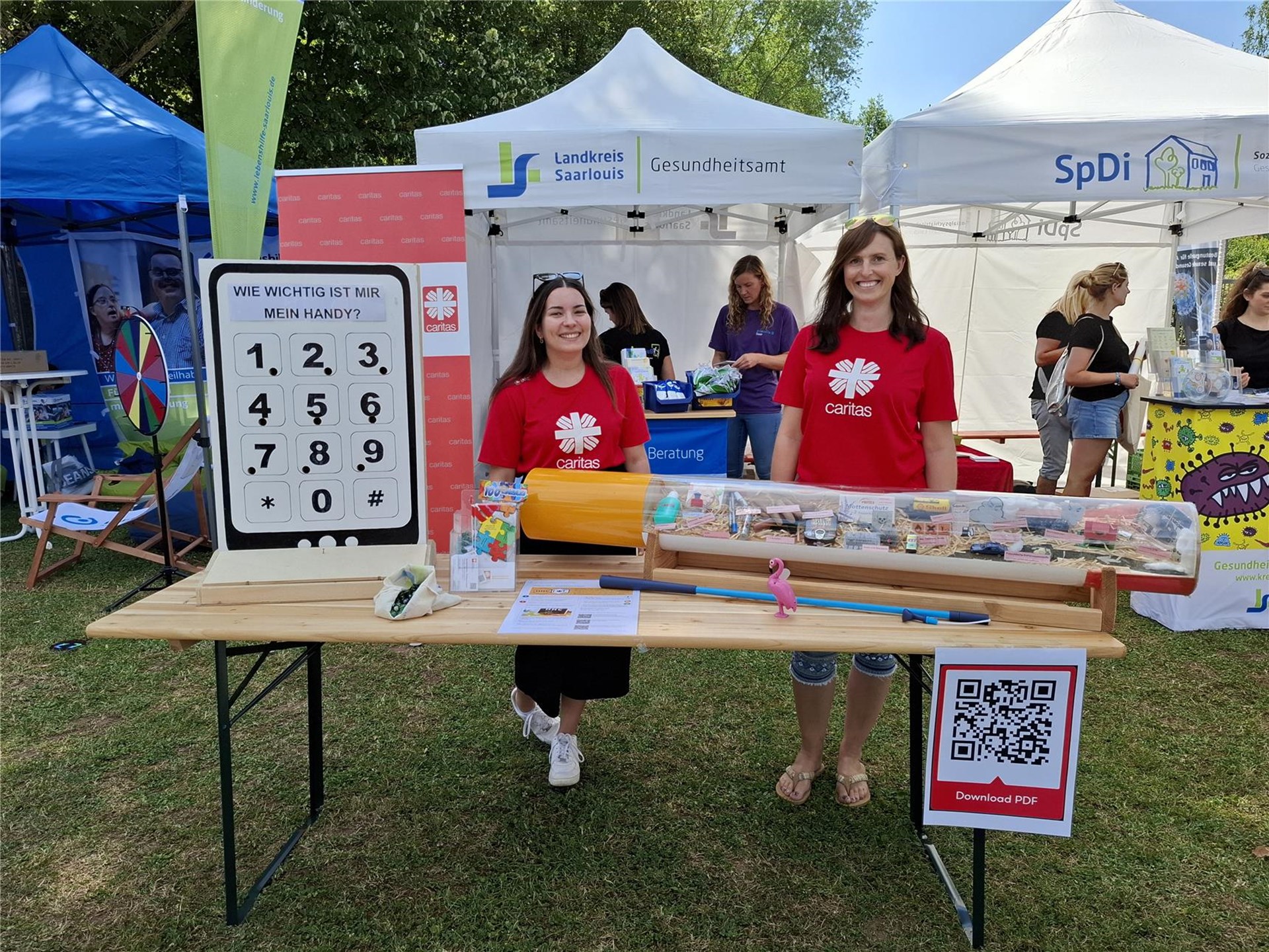 Zwei Frauen in roten Caritas-T-Shirts stehen an einem Infostand auf einem Freiluftfest mit interaktiven Materialien und Plakat zur Handynutzung.