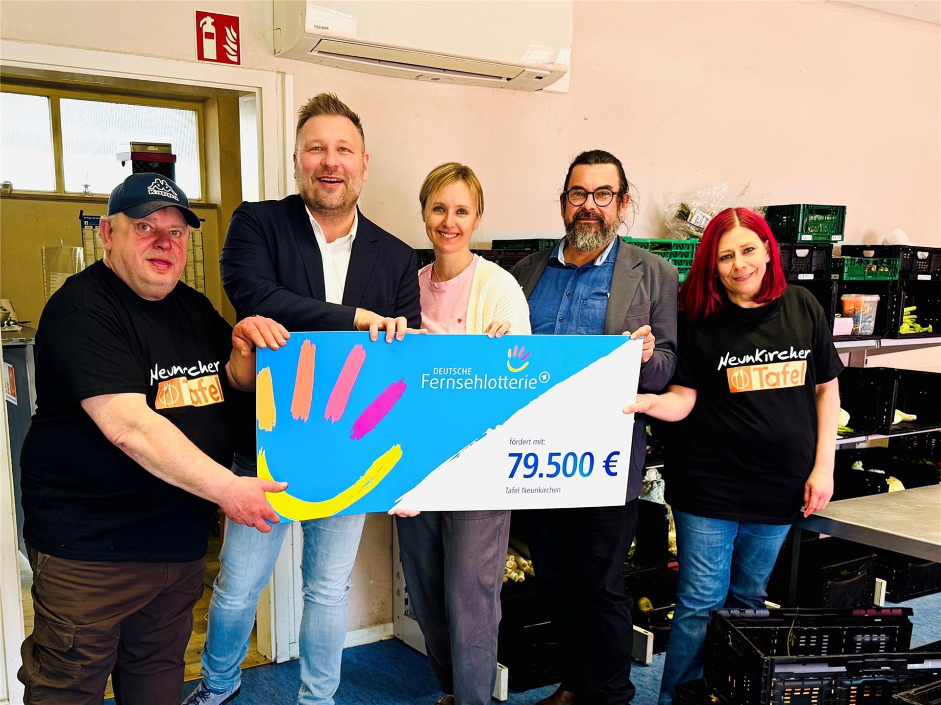 5 Menschen stehen hinter in den Räumen der Neunkircher Tafel und halten einen Scheck der deutschen Fernsehlotterie in Höhe von 79.500 € in den Händen.