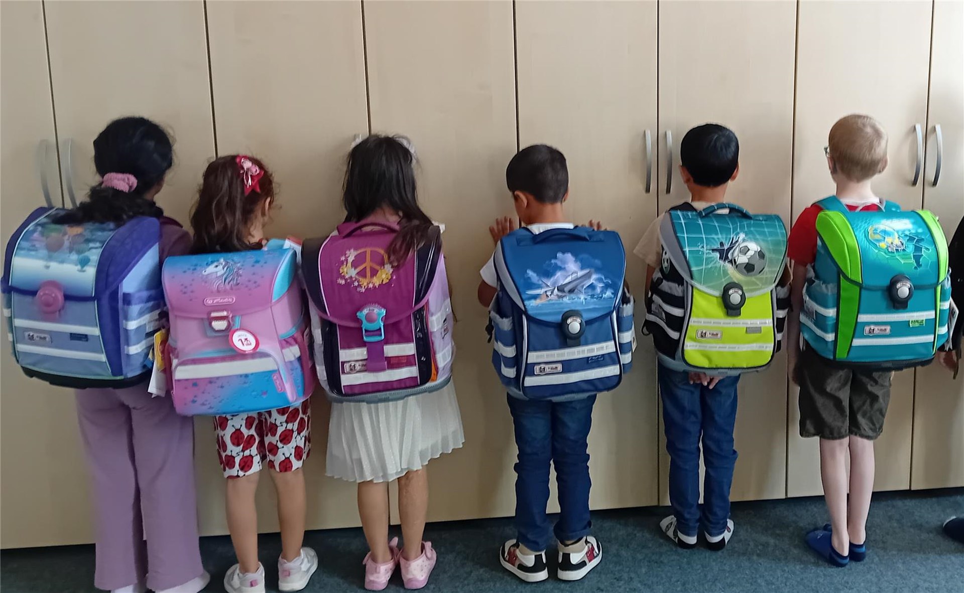 6 Kinder stehen mit dem Rücken zur Kamera. Sie tragen neue Schulranzen.