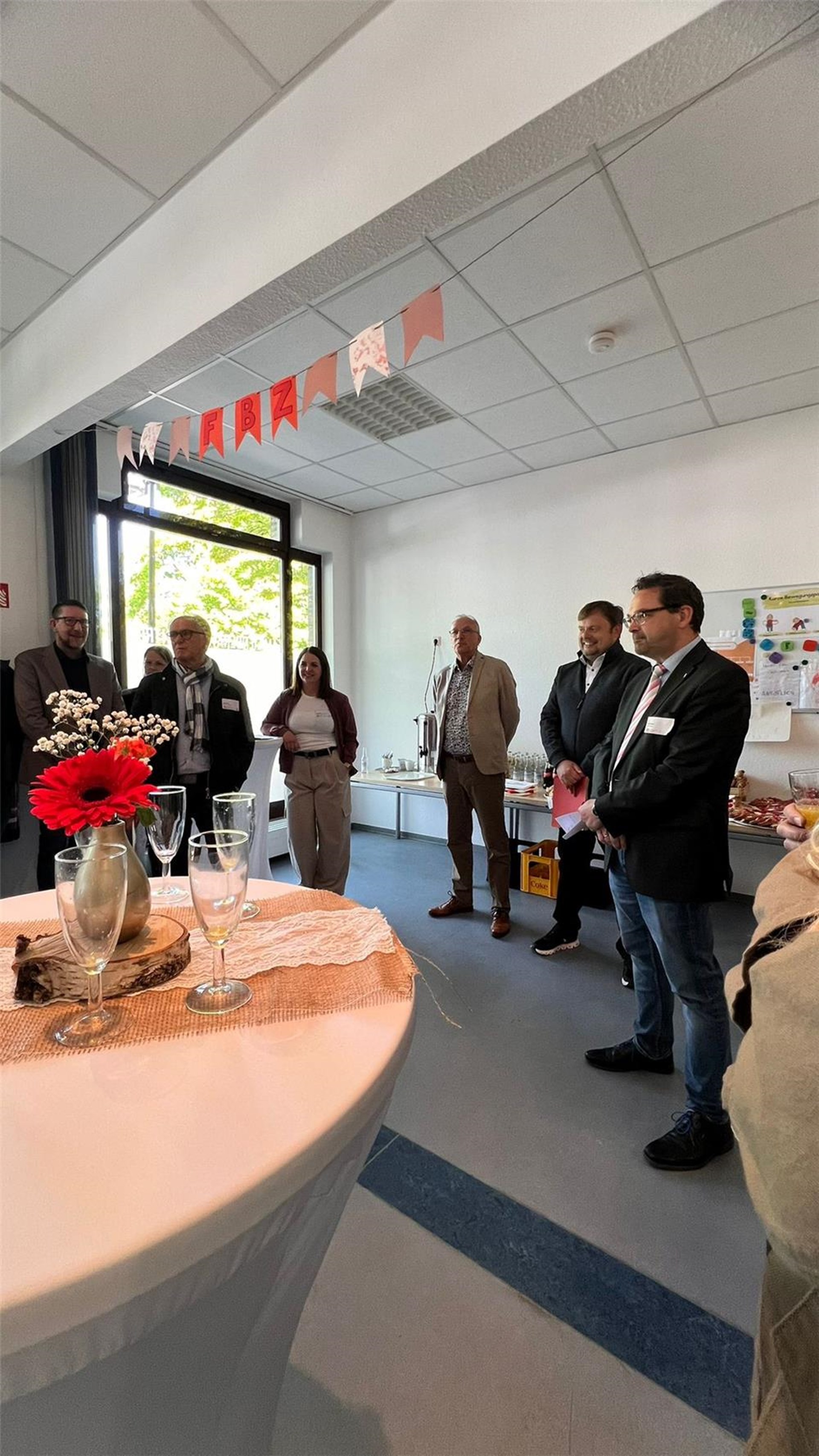 Landrat Udo Recktenwald, Caritasdirektor Dirk Schmitt, sowie Bürgermeister Volker Weber stehen in den Räumen des FBZ Marpingen. Es ist eine Girlande zusehen, sowie ein Stehtisch mit Sektgläsern. 