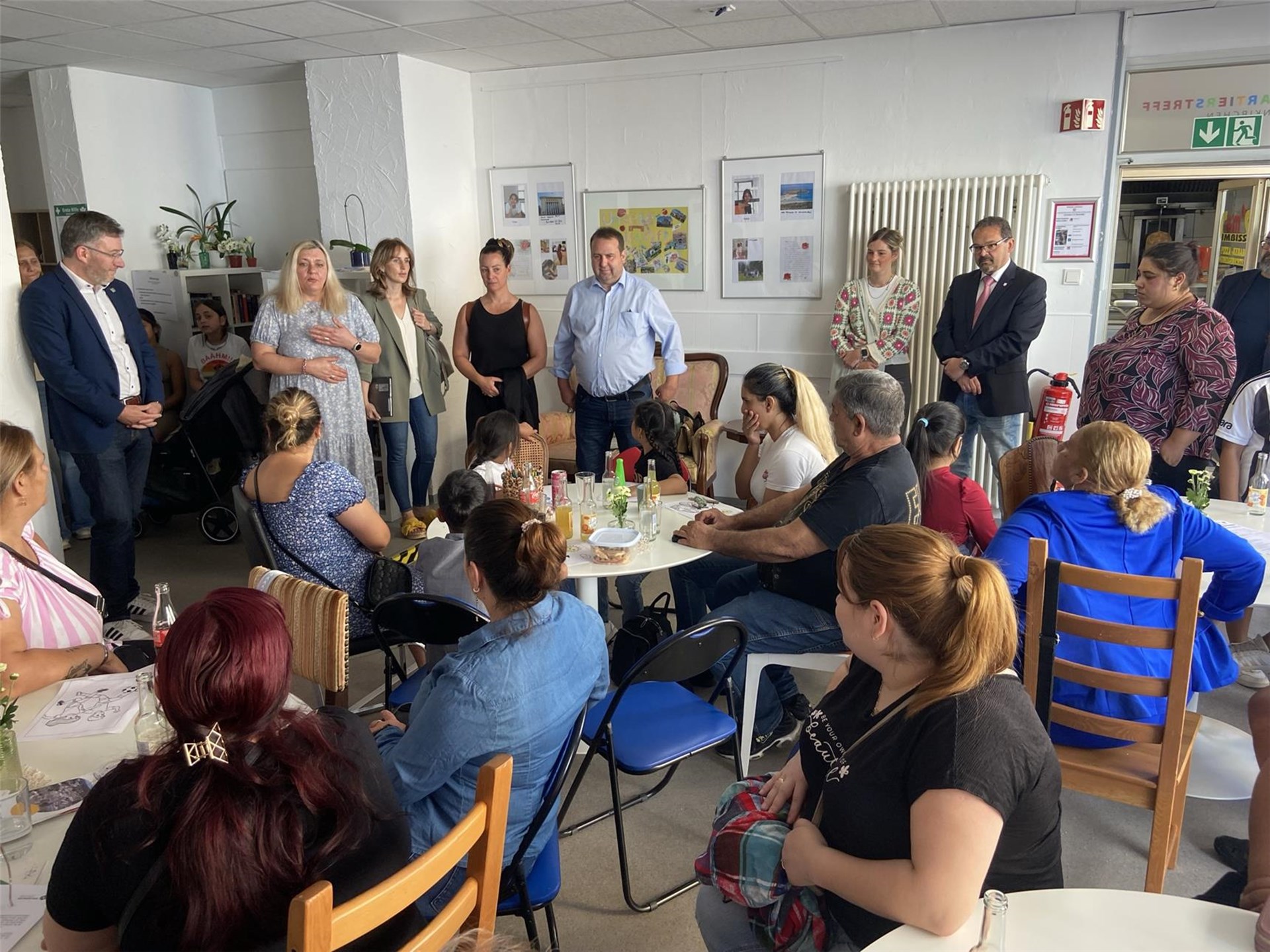 Mehrere Menschen sitzen in einem Gemeinschaftsraum an Tischen, während eine Gruppe von Personen vorne steht und spricht. Eine dieser Personen ist Minister Dr. Magnus Jung. 