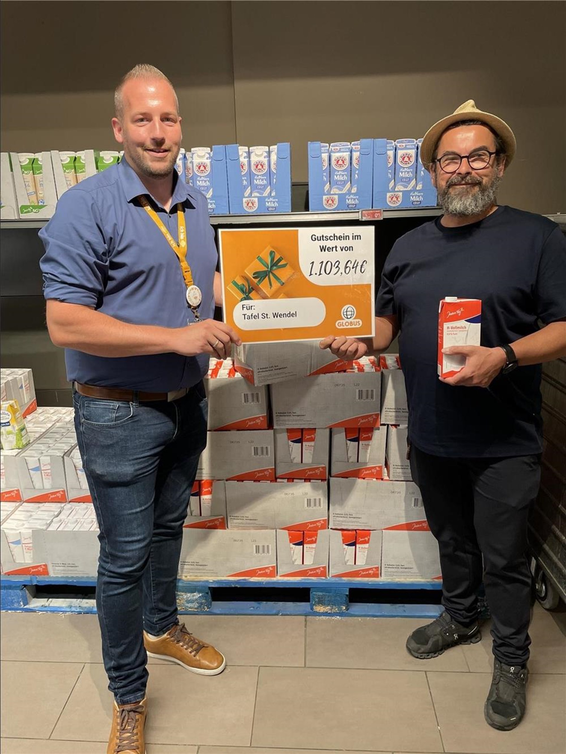 2 Menschen halten einen symbolischen Spendenscheck gemeinsam hoch. Darauf steht ein Betrag von 1.103,64 €. Der Mann auf der rechten Seite hält außerdem eine Tüte Milch.