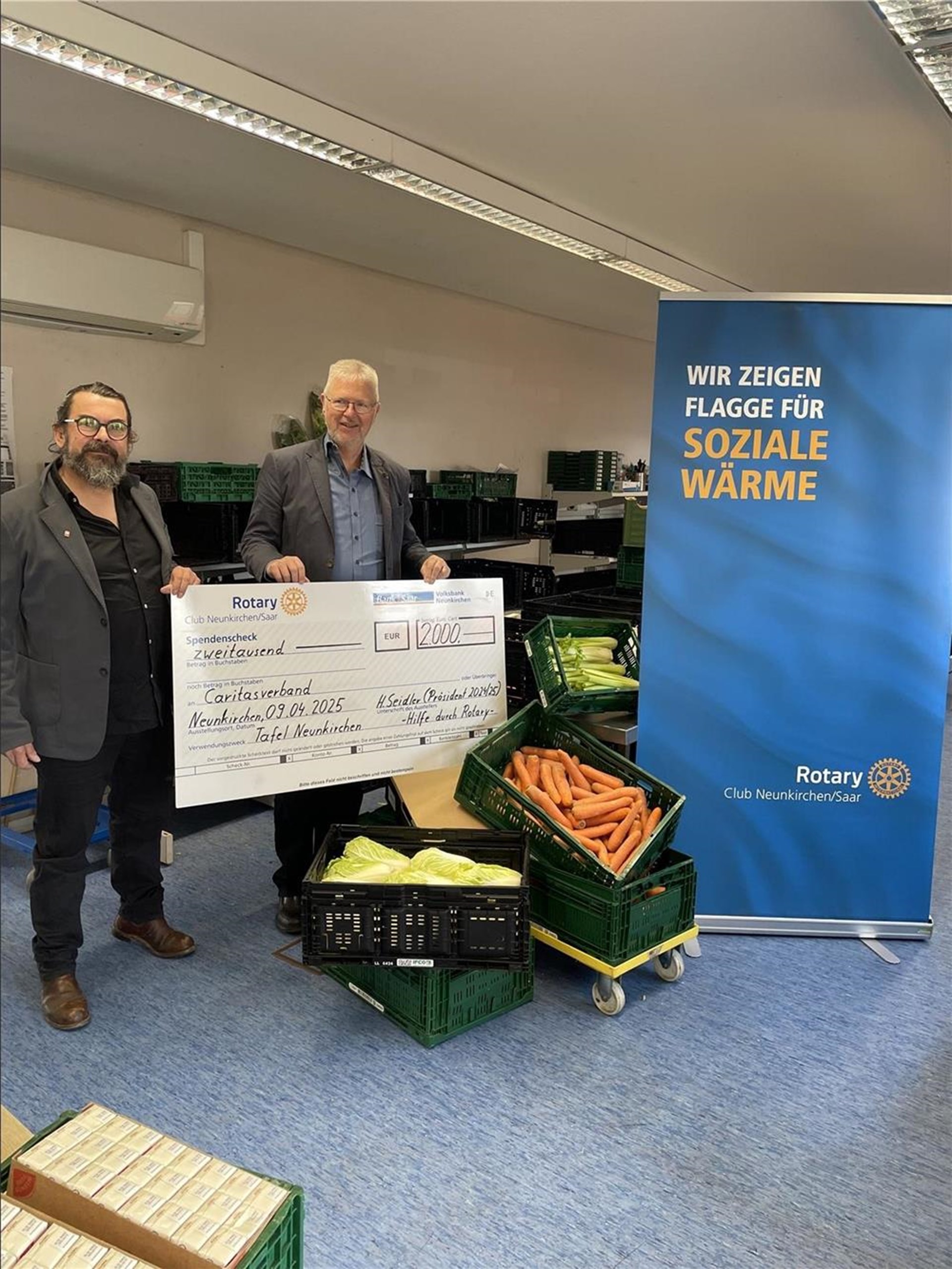 Thomas Mördorf und Harald Seidler halten gemeinsam einen symbolischen Scheck über 2.000 €. Sie befinden sich in den Räumen der Tafel.  Im Hintergrund ist ein Roll-up des Rotary-Clubs zu sehen