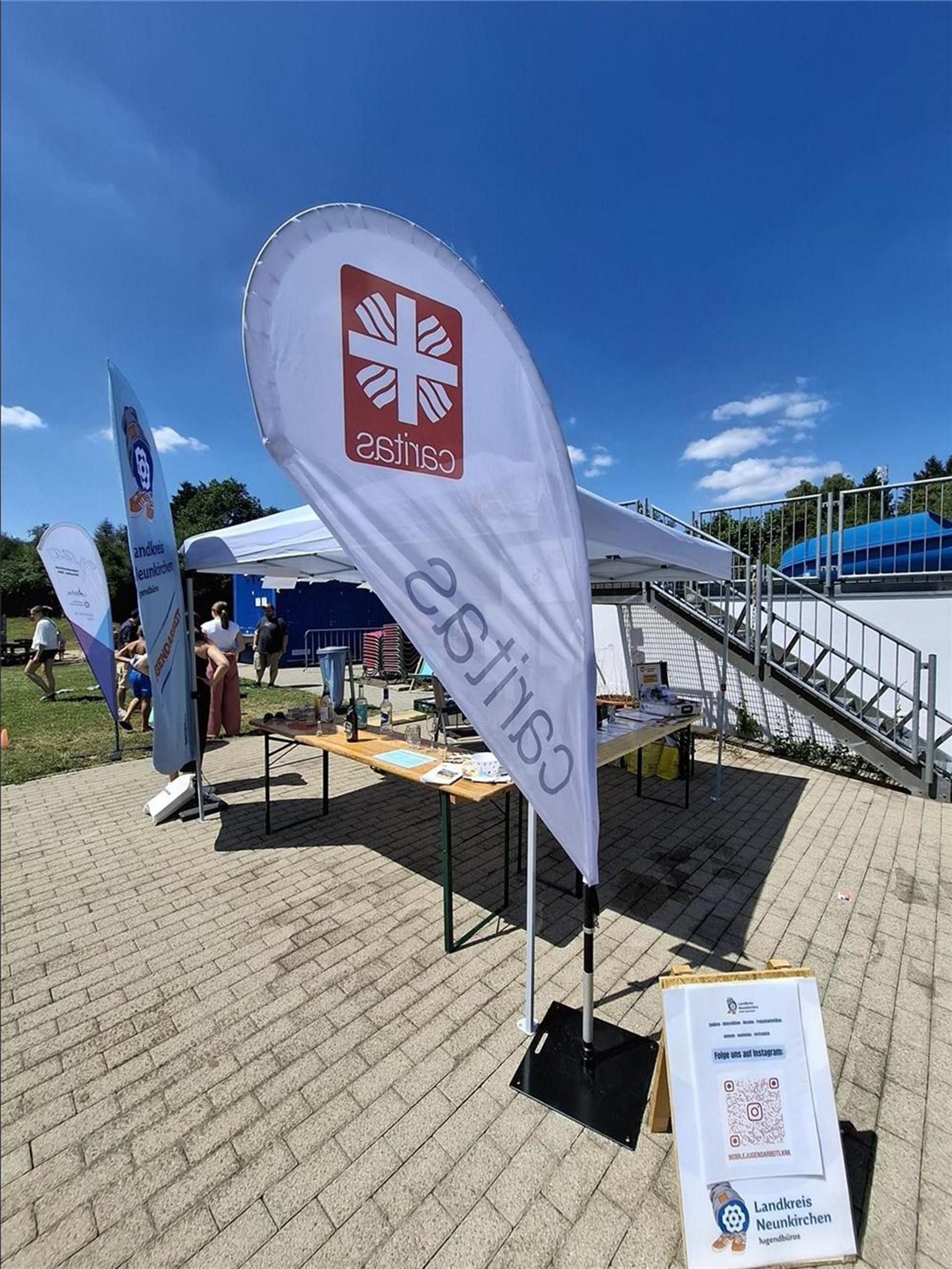 Ein Informationsstand der Caritas mit großen Fahnen und Flyern steht im Freien bei sonnigem Wetter, daneben ein Aufsteller des Landkreises Neunkirchen.
