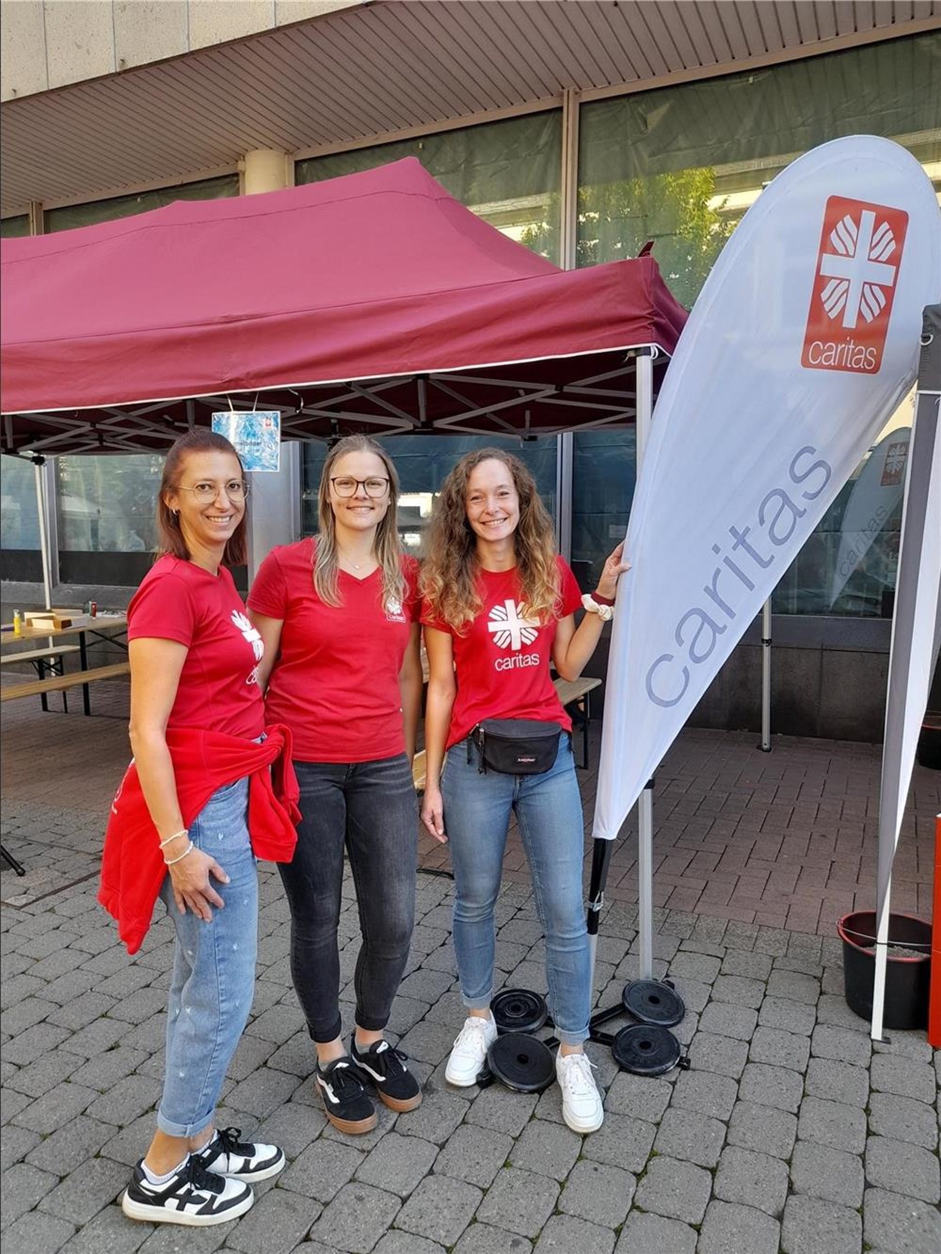 3 Mitarbeiterinnen der Caritas stehen mit roten Shirts neben einer weißen Beachflag mit Caritas- Schriftzug